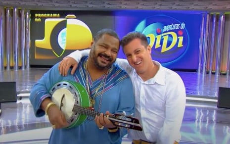 O cantor Arlindo Cruz e o apresentador Luciano Huck na vinheta Samba da Globalização de 2012, da Globo, com um telão ao fundo que mostra o Programa do Jô e As Aventuras de Didi