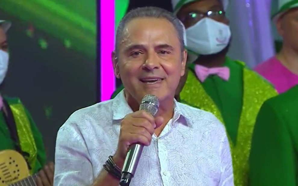 Luís Roberto com uma blusa azul e microfone na mão no programa Seleção do Samba, da Globo
