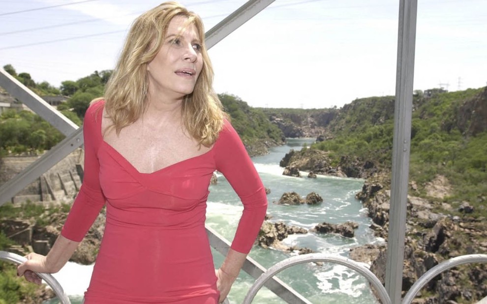 A atriz Renata Sorrah com um vestido vermelho, decotado, com expressão de preocupação em uma ponte de metal sobre o rio São Francisco em cena de Senhora do Destino