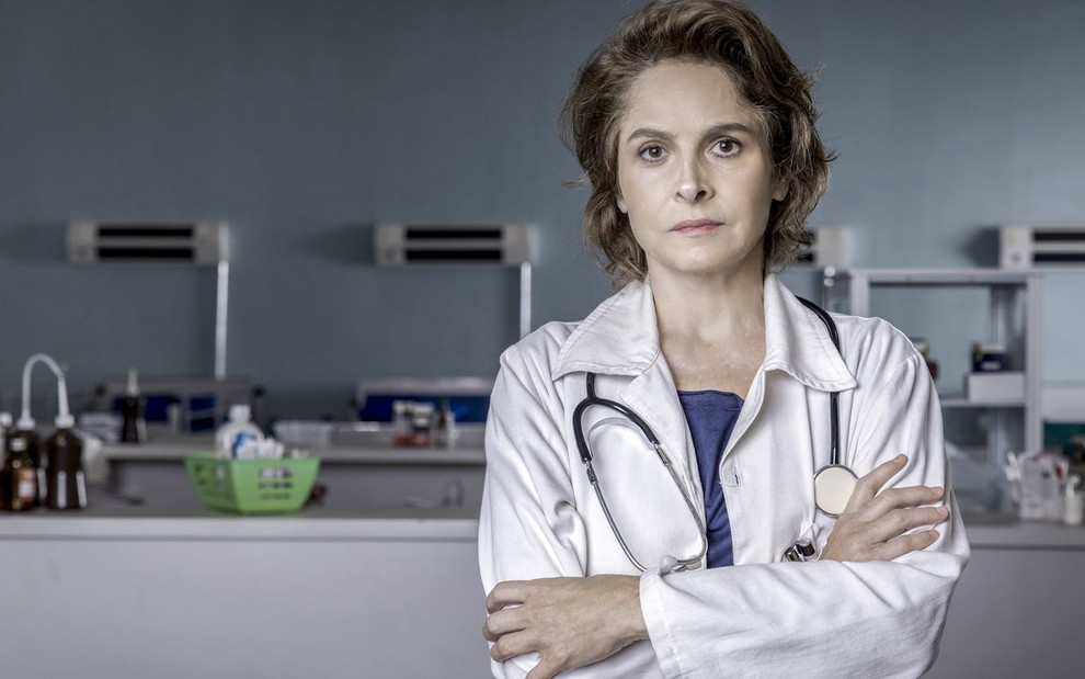 A atriz Drica Moraes usa jaleco branco, com estetoscópio pendurado no pescoço, em cenário de hospital de Sob Pressão