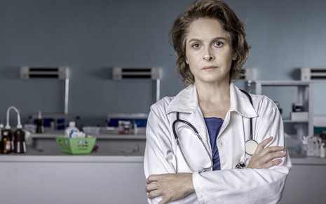 A atriz Drica Moraes usa jaleco branco, com estetoscópio pendurado no pescoço, em cenário de hospital de Sob Pressão