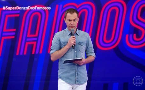O apresentador Tiago Leifert usa camisa jeans e faz cara de bravo com um microfone na mão durante gravação da Super Dança dos Famosos