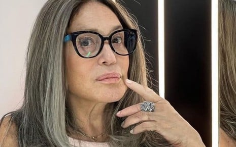 Susana Vieira usa uma blusa branca e um óculos de aro preto. Ela encara a câmera, séria, e posa um dedo perto da boca.