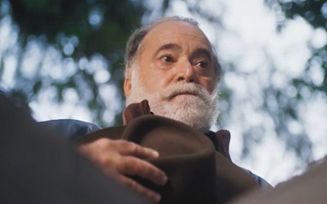 O ator Tony Ramos segura o chapéu no peito e tem expressão boçal em cena da novela Terra e Paixão