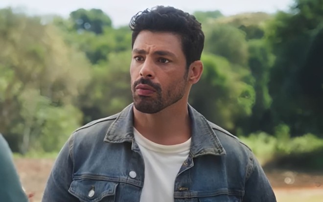 O ator Cauã Reymond em cena como Caio em Terra e Paixão, com expressão séria, camisa de manga longa cinza e camiseta branca