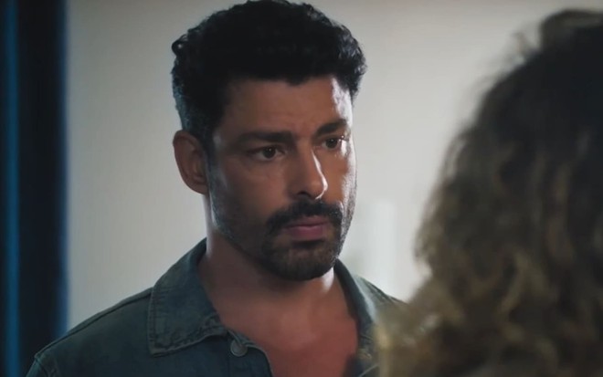 Cauã Reymond caracterizado como Caio; ele usa uma regata branca e está sério em cena de Terra e Paixão