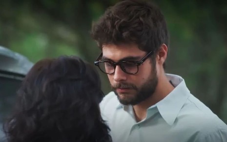 Rafael Vitti caracterizado como Hélio; ele tem o cabelo bem aparado e usa uma blusa polo branca enquanto encara Debora Ozório --de costas para a câmeéji