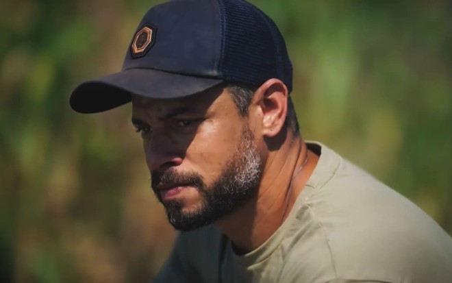 Paulo Lessa tem os cabelos curto e usa uma regata bege e uma camisa verde; ele sorri em cena de Terra e Paixão