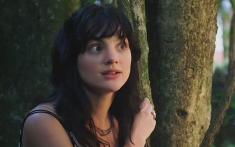 A atriz Debora Ozório em cena de Terra e Paixão, com expressão de medo, atrás de árvore