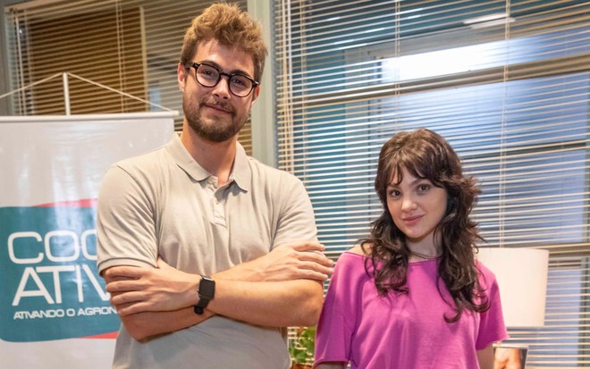 Rafa Vitti, o Hélio, tem os cabelos curtos e barba longa e veste uma camisete bege. Já Debora Ozório, a Petra, tem os cabelos escuros, longos e soltos, e veste uma camisa rosa