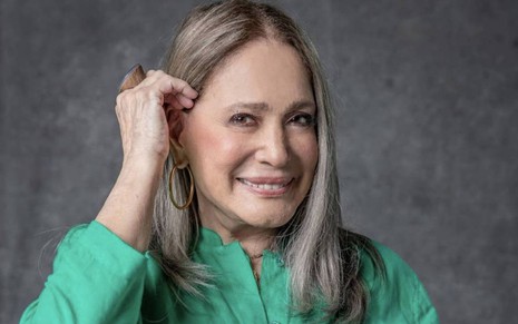 Susana Vieira usa uma blusa verde e um brinco dourado. Ela encara a câmera, sorridente, com uma das mãos apoiadas nos cabelos lisos e grisalhos