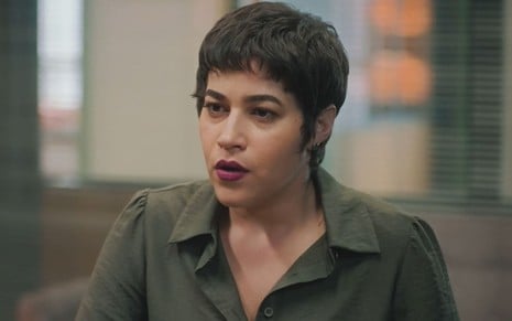 Yasmin Gomlevsky caracterizada como Isabel; ela tem os cabelos pretos curtos e usa uma jaqueta de sarja bege em cena de Terra e Paixão. O semblante exprime tensão.