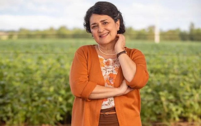 A atriz Inez Viana está com casaco laranja, blusa estampada e posa em um campo que serve de cenário para Terra e Paixão