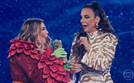 A cantora Rosana está com fantasia vermelha, luvas verdes e microfone na mão enquanto fala com Ivete Sangalo no The Masked Singer Brasil