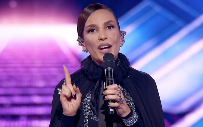 Ivete Sangalo em The Masked Singer Brasil: cantora usa roupa preta, segura microfone de mão e levanta o dedo em riste
