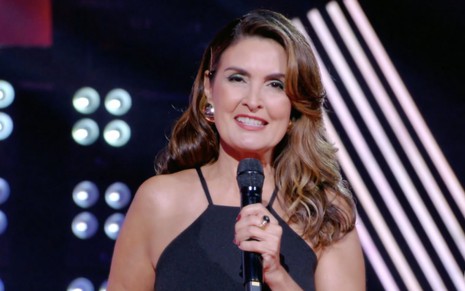 Fátima Bernardes segura um microfone com a mão direita no palco do The Voice Kids