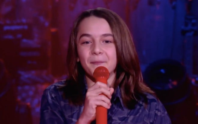 O participante do The Voice Kids Henrique Bonadio segura um microfone vermelho e sorri durante o programa