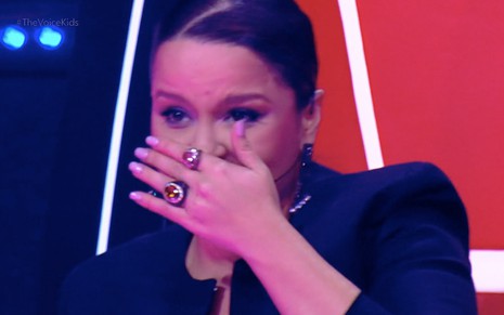 A cantora Maiara leva a mão ao rosto para limpar as lágrimas na cadeira do The Voice Kids