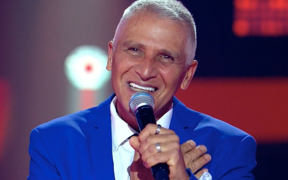 O cantor Maurício Gasperini no palco do The Voice+ com um microfone na mão direita e um terno azul com blusa branca
