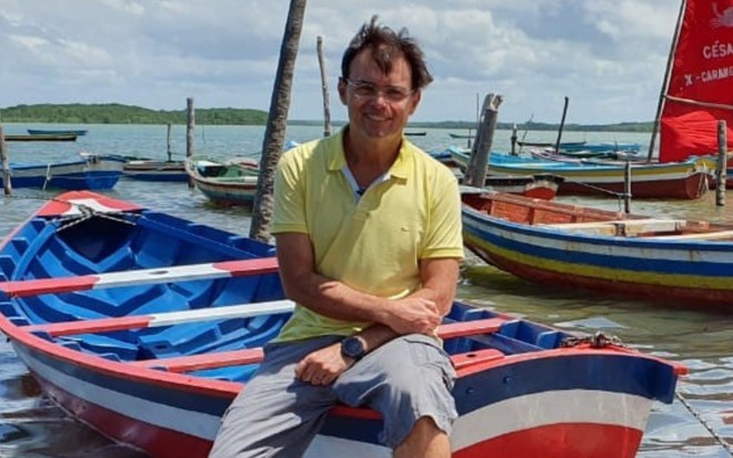 O repórter Tino Marcos está sentado em um barco; ele usa camiseta amarela e está sorridente