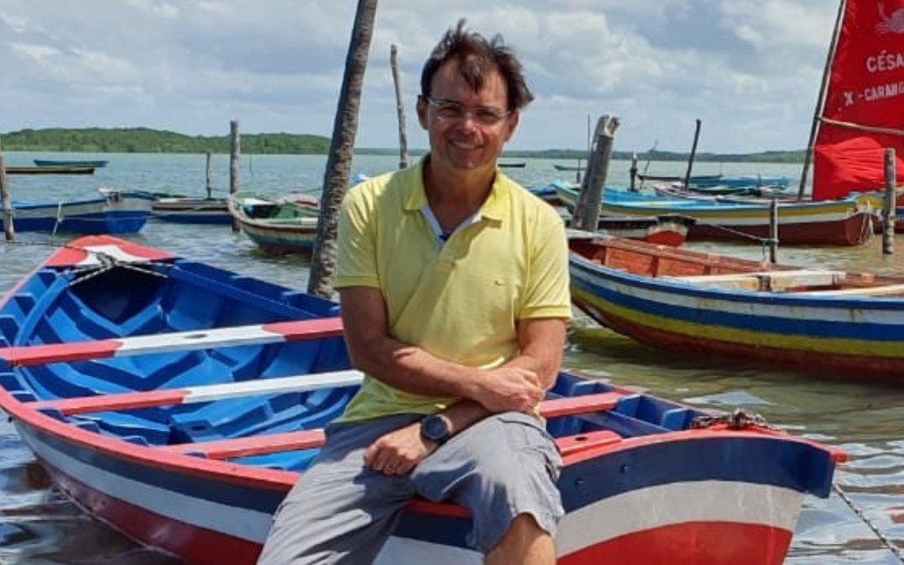 O repórter Tino Marcos está sentado em um barco; ele usa camiseta amarela e está sorridente