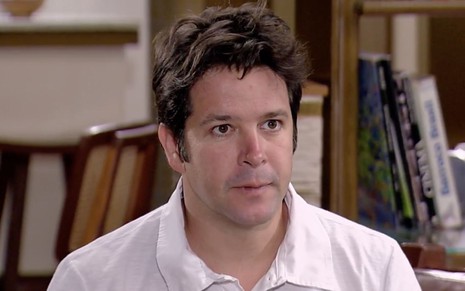 Murilo Benício caracterizado como Ariclenes em cena de Ti Ti Ti: de camiseta branca, rapaz está sentado e olha com choque para alguém fora do quadro