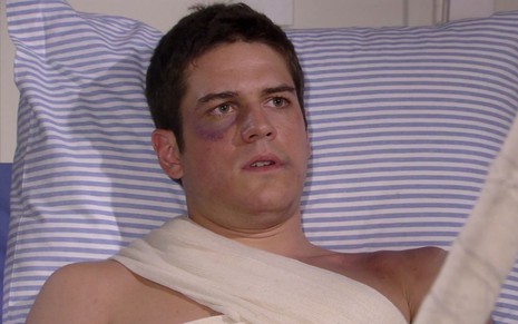 Marco Pigossi em cena de Ti Ti Ti: caracterizado como Pedro, ator está deitado em cama de hospital, com olho roxo e braço enfaixado
