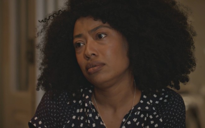 Mariana Nunes tem a expressão indignada em cena de Travessia; ela usa os cabelos crespos longos e veste uma blusa florida