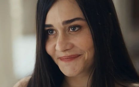 Alessandra Negrini caracterizada como Guida; ela usa uma camiseta preta e dá um sorriso cúmplice em cena de Travessia