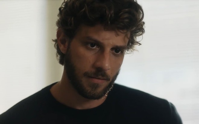 Chay Suede, caracterizado como Ari, usa uma camiseta preta e os cabelos cacheados, curtos; a expressão está transtornada