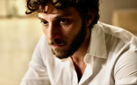O ator Chay Suede usa camisa branca e olha para a frente com expressão tensa em cena da novela Travessia como Ari