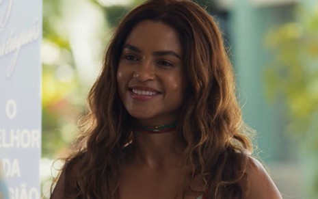 Lucy Alves, caracterizada como Brisa, sorri e usa uma blusa florida em cena de Travessia