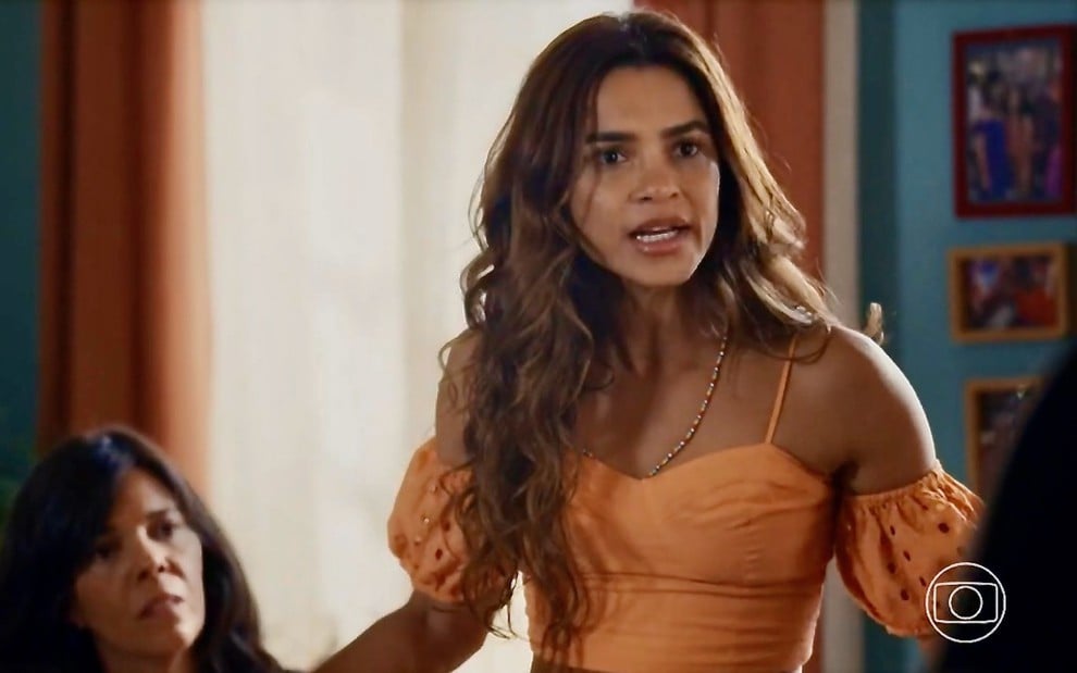 A atriz Lucy Alves em cena da novela Travessia com um top laranja e expressão de brava