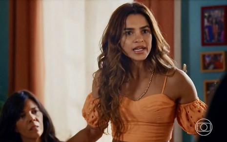 A atriz Lucy Alves em cena da novela Travessia com um top laranja e expressão de brava