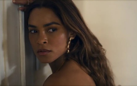 Lucy Alves está encostada em uma porta com os cabelos soltos em cena da novela Travessia, da Globo
