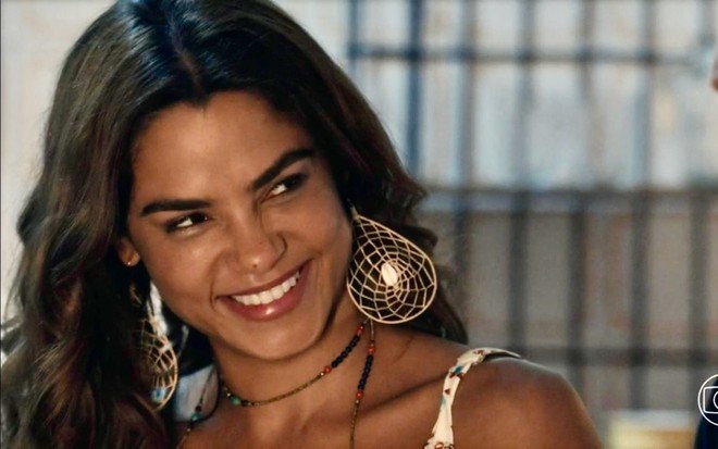 A atriz Lucy Alves sorri em cena da novela Travessia caracterizada como Brisa, com cabelos soltos, brincos grandes e blusa de alcinha