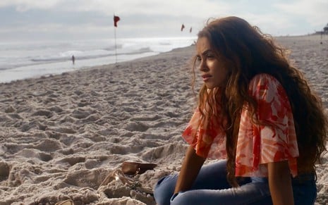A atriz Lucy Alves usa blusa estampada e está com cabelos soltos sentada na areia de uma praia em cena da novela Travessia