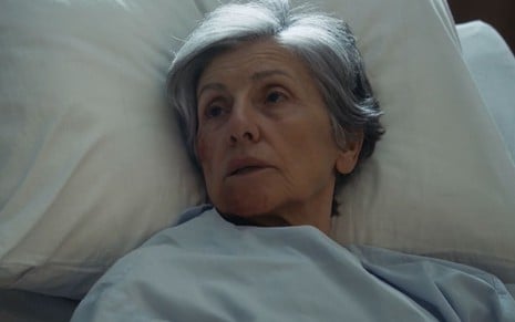 Cassia Kis caracterizada como Cidália; ela está deitada num leito de hospital e tem a boca aberta em cena de Travessia