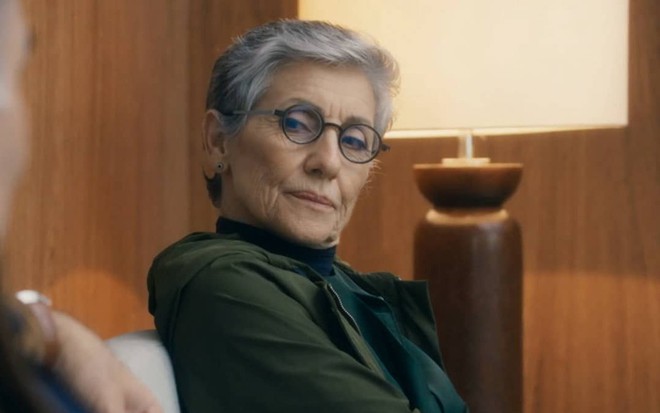A atriz Cassia Kis olha para a frente em cena de Travessia na qual usa óculos e faz uma expressão de desagrado como Cidália