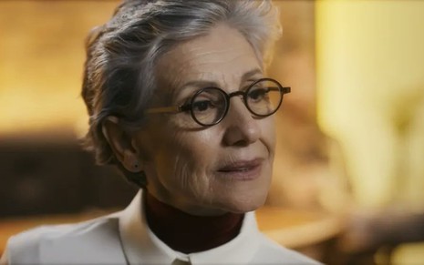 A atriz Cassia Kis usa óculos e camisa clara caracterizada como Cidália em cena da novela Travessia, da Globo
