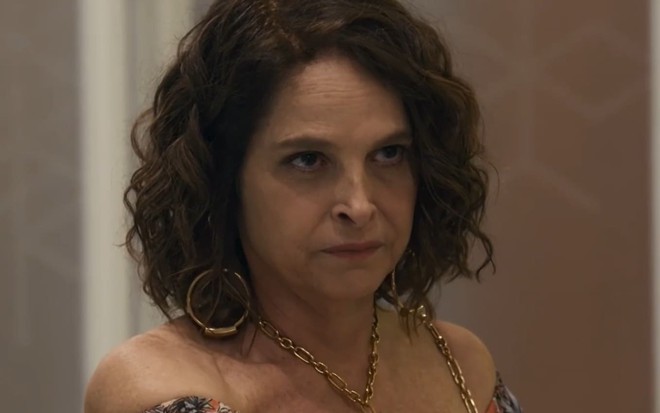 Drica Moraes caracterizada como Núbia em Travessia; ela usa um vestido laranja e tem o semblante sério em cena da novela