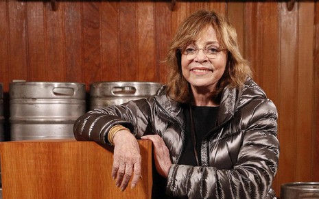 A autora Gloria Perez posa em cenário de bar da novela Travessia; ela usa um casaco prateado, blusa preta e óculos