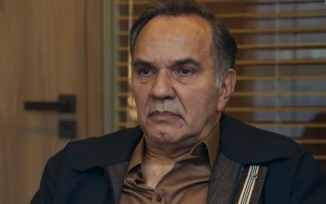 Humberto Martins caracterizado como Guerra em Travessia; ele tem o semblante firme e usa uma camisa branca e uma jaqueta bege em cena da novela