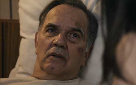 O ator Humberto Martins está em uma cama com maquiagem de hematomas no rosto em gravação de cena da novela Travessia