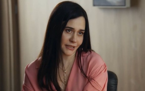 Alessandra Negrini caracterizada como Guida em Travessia; ela usa uma camisa rosa e colares dourados e tem o semblante transtornado em cena da novela
