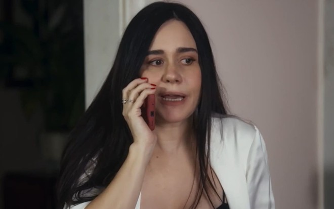 Alessandra Negrini caracterizada como Guida em Travessia; ela usa uma camisa rosa e colares dourados e tem o semblante transtornado em cena da novela