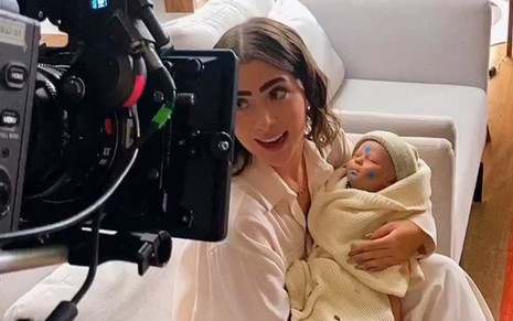 Jade Picon está diante de uma câmera com um boneco bebê no lugar de um recém-nascido em gravação de Travessia