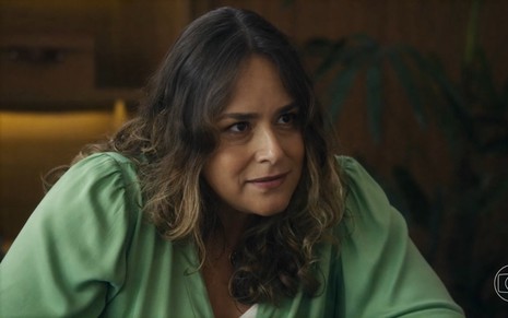 A atriz Tabata Contri usa blusa verde e encara alguém, que está fora da imagem, em cena da novela Travessia