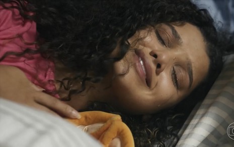 A atriz Danielle Olímpia está chorando, deitada em uma cama, em cena da novela Travessia como a personagem Karina