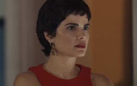 Vanessa Giácomo, caracterizada como Leonor, dá um meio sorriso em cena de Travessia; ela usa os cabelos bem curtos e veste uma regata vermelha